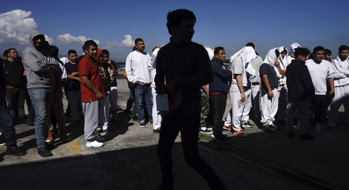 Grupo de personas de origen cubano y venezolano con maletas y bolsas sentadas afuera de un albergue migrante en Tapachula, Chiapas, México, con expresiones de agotamiento e incertidumbre, mientras un letrero del Instituto Nacional de Migración de México se ve al fondo.