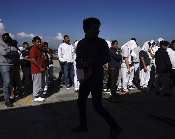Grupo de personas de origen cubano y venezolano con maletas y bolsas sentadas afuera de un albergue migrante en Tapachula, Chiapas, México, con expresiones de agotamiento e incertidumbre, mientras un letrero del Instituto Nacional de Migración de México se ve al fondo.